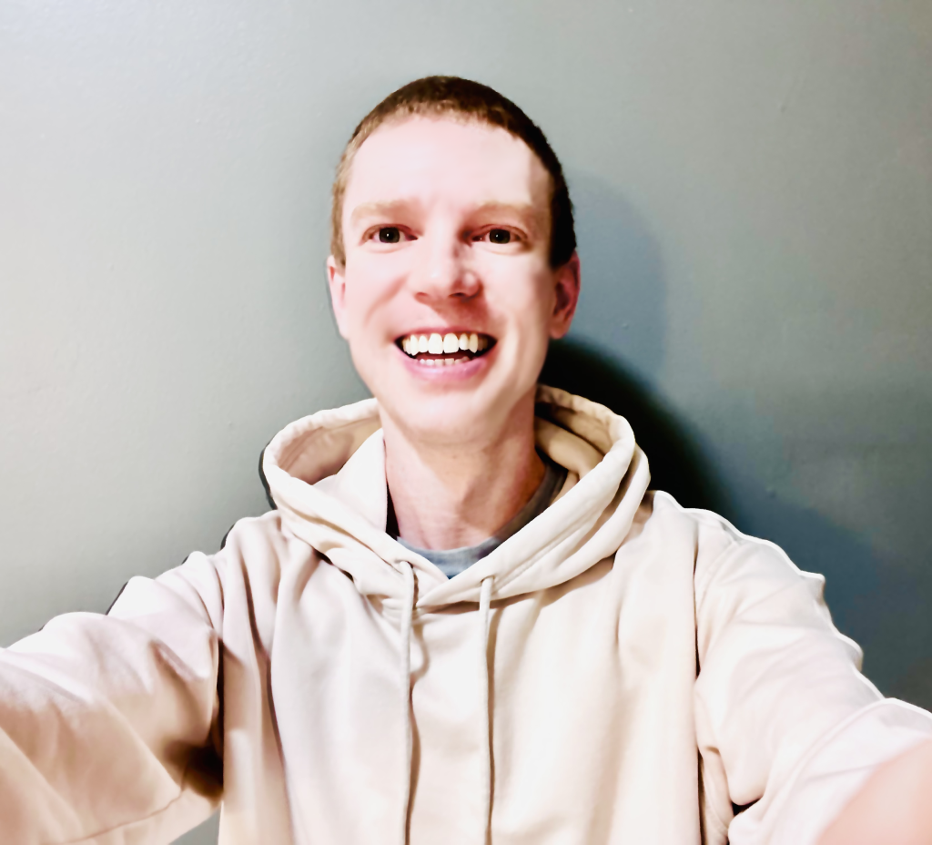 Logan W Reynolds, smiling and wearing a light-colored hoodie, taking a selfie against a plain gray background.